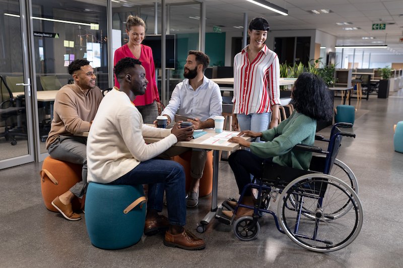 A diverse group of colleagues, including a wheelchair user, collaborate and converse in a modern office setting, illustrating social and community participation.