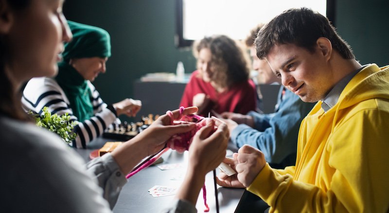 A group of people, including individuals with disabilities, engage in various activities like knitting and playing cards, exemplifying social and community participation.