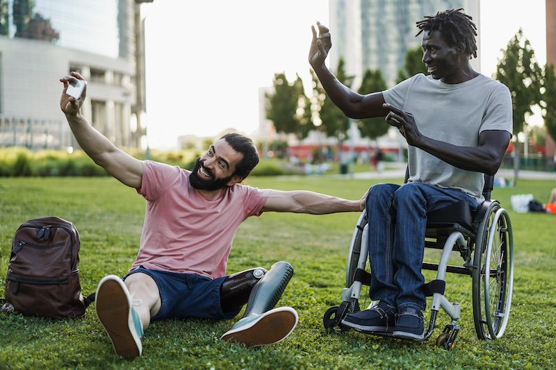 Two friends, one with a prosthetic leg and the other in a wheelchair, enjoy a joyful moment in an urban park, highlighting social and community participation.