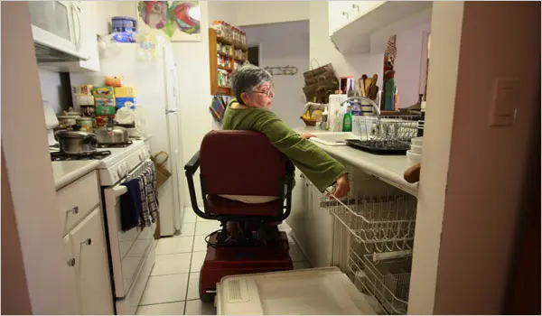 A person using a wheelchair is sitting in a kitchen and reaching into a dishwasher, demonstrating daily activities NDIS. The kitchen is equipped with various appliances and items, reflecting a well-organized and functional space.