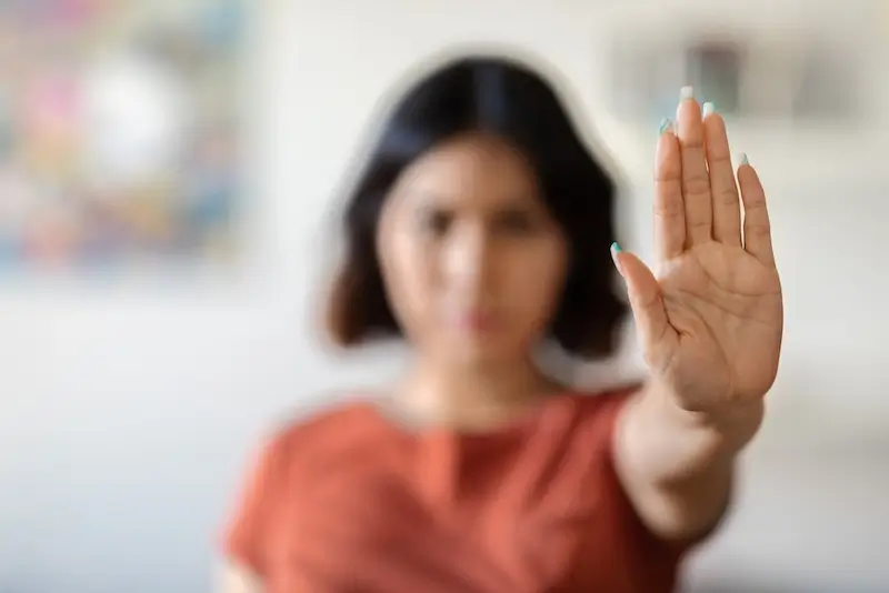 A blurred image of a woman in an orange shirt extending her hand towards the camera, palm facing forward in a gesture of "stop." The photo symbolises a stand against domestic violence.