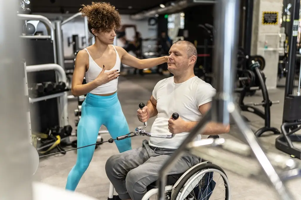 A man in a wheelchair exercises with the guidance of a trainer, demonstrating the principles of exercise physiology in an adaptive fitness setting.