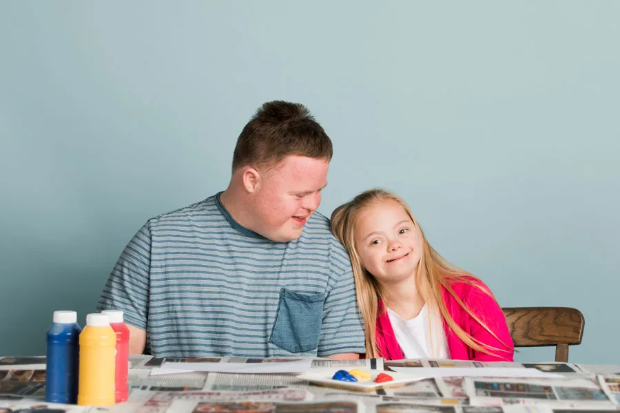 A boy and a girl with Down syndrome sitting at a table covered with art supplies, demonstrating Positive Behaviour Support.