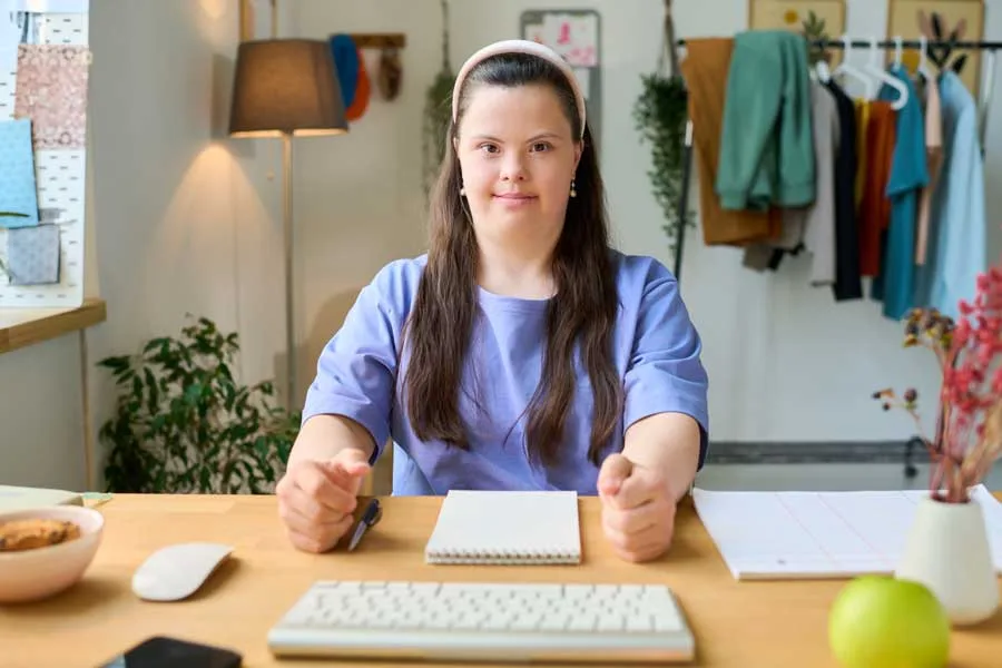 Young woman with Down syndrome sitting at a desk in a well-organized room, illustrating her productive and independent lifestyle with NDIS Supported Independent Living Program.