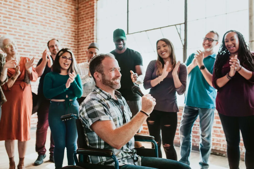 A man in a wheelchair speaking into a microphone while a diverse group of people claps and smiles, illustrating community participation.