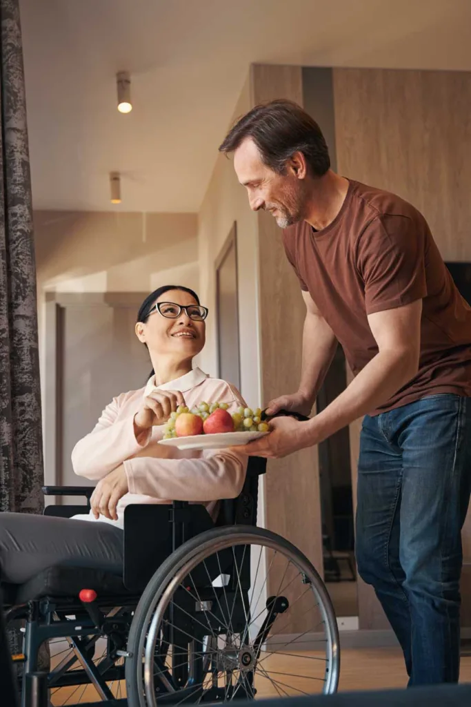 A man serving a plate of fruits to a woman in a wheelchair, emphasizing the supportive role of a carer.