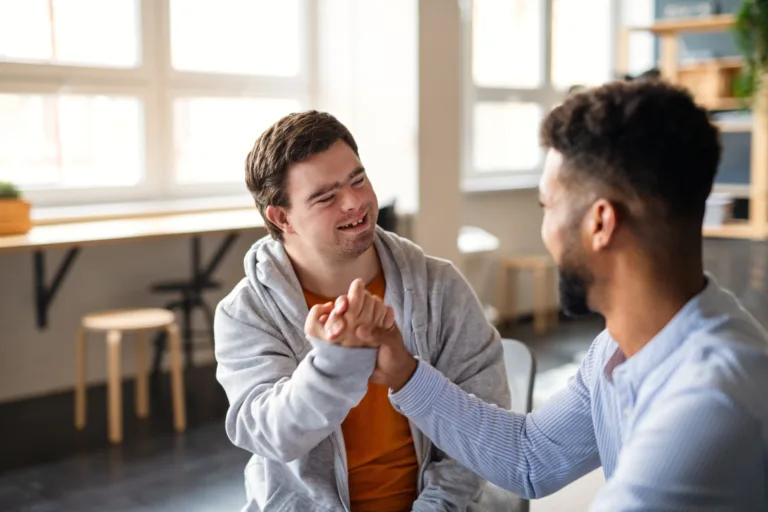 Two men, one with Down syndrome, smiling and shaking hands in a bright room with large windows.