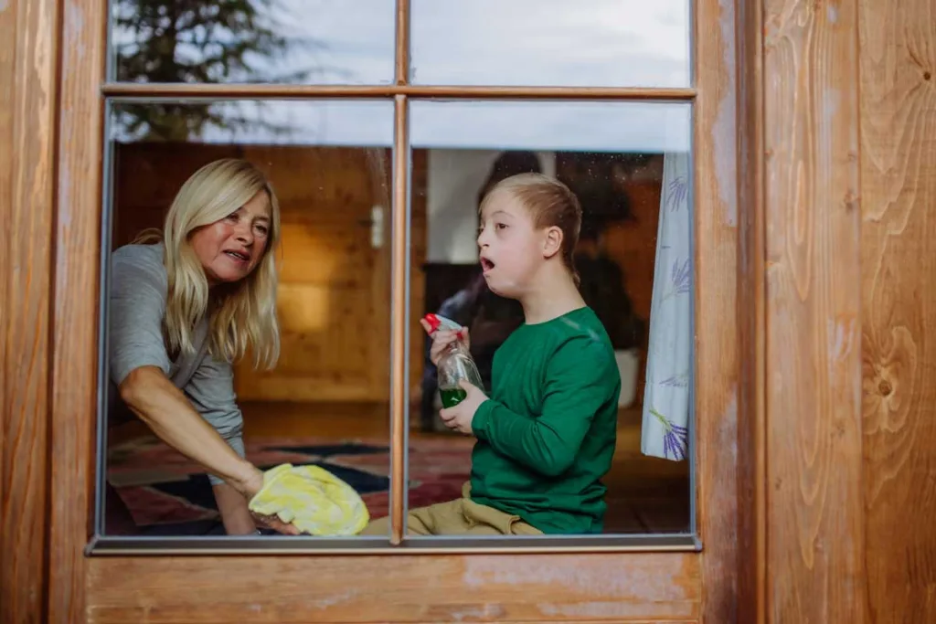 A mother and her young son with Down syndrome engaging in household cleaning together through a window, illustrating an NDIS-supported activity that promotes independence and life skills.