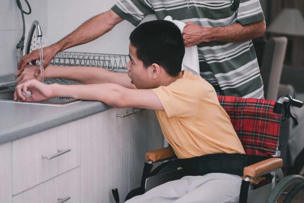 A young boy in a wheelchair washing dishes at the kitchen sink with assistance from an elderly man, highlighting NDIS household tasks.