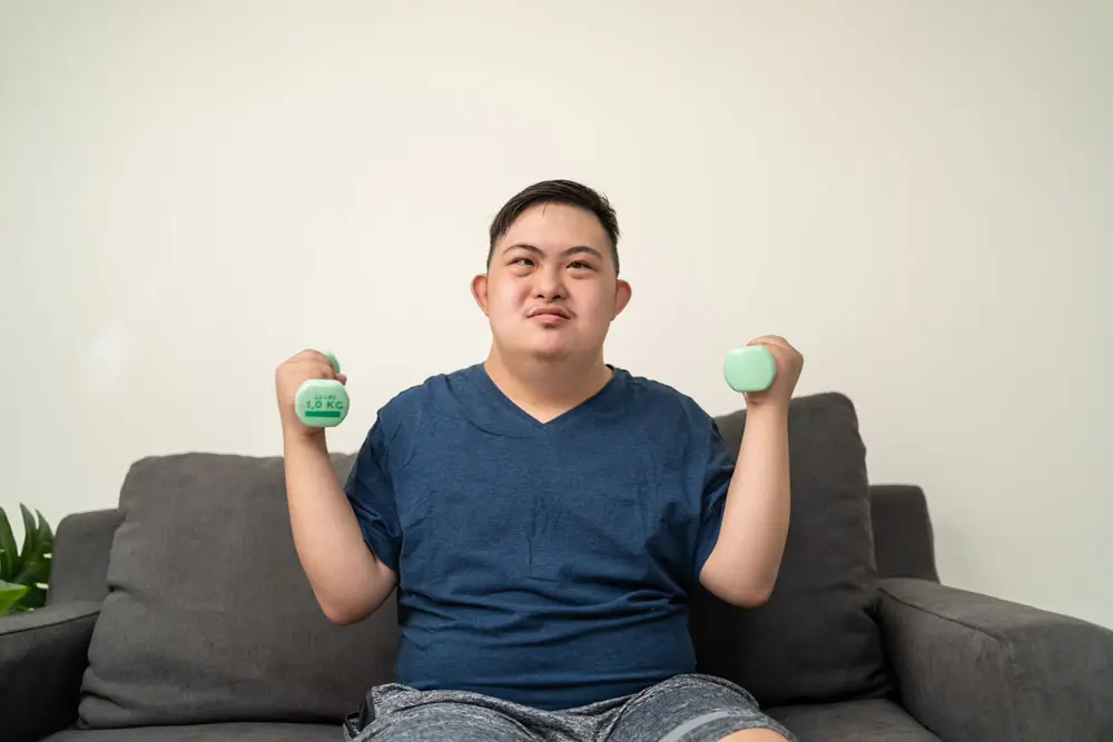 A young man with Down syndrome engages in a home workout, lifting weights while seated on a couch, as part of his NDIS exercise physiology program to enhance strength and coordination.