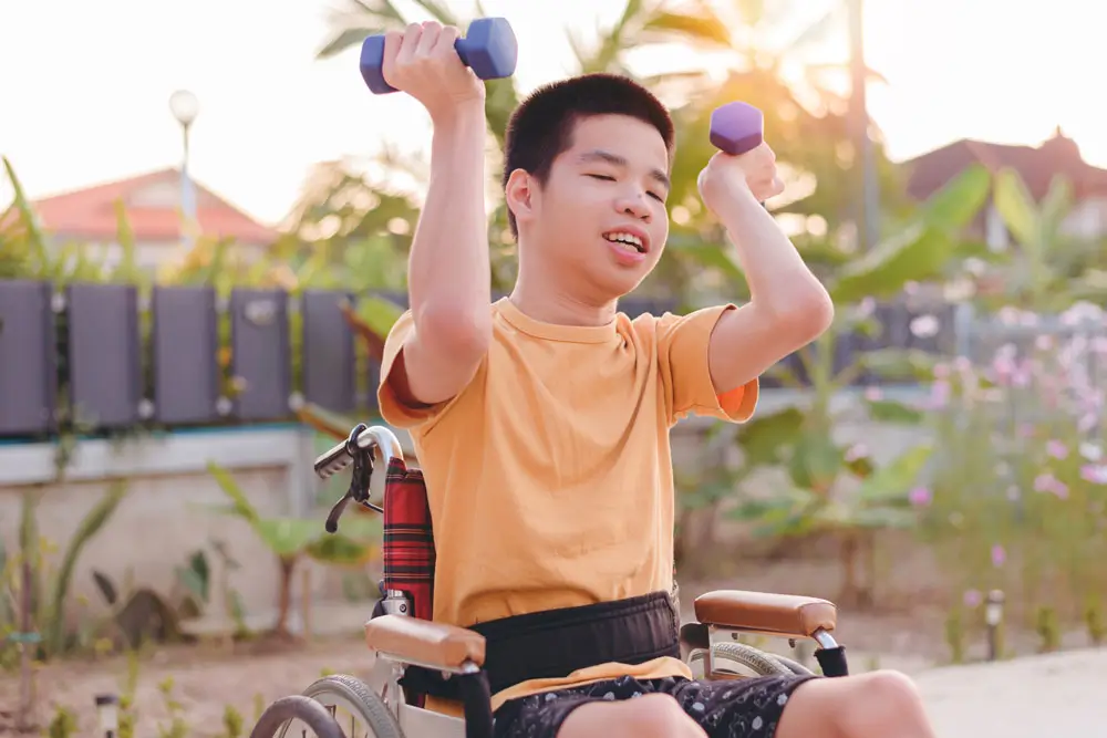 An young boy in a wheelchair performing strength training with dumbbells outdoors, as part of his NDIS daily activities to promote physical health and well-being.