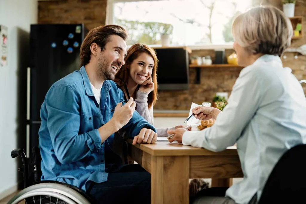 A man in a wheelchair and a woman smiling warmly during a meeting with a support coordination professional at a homely kitchen table.