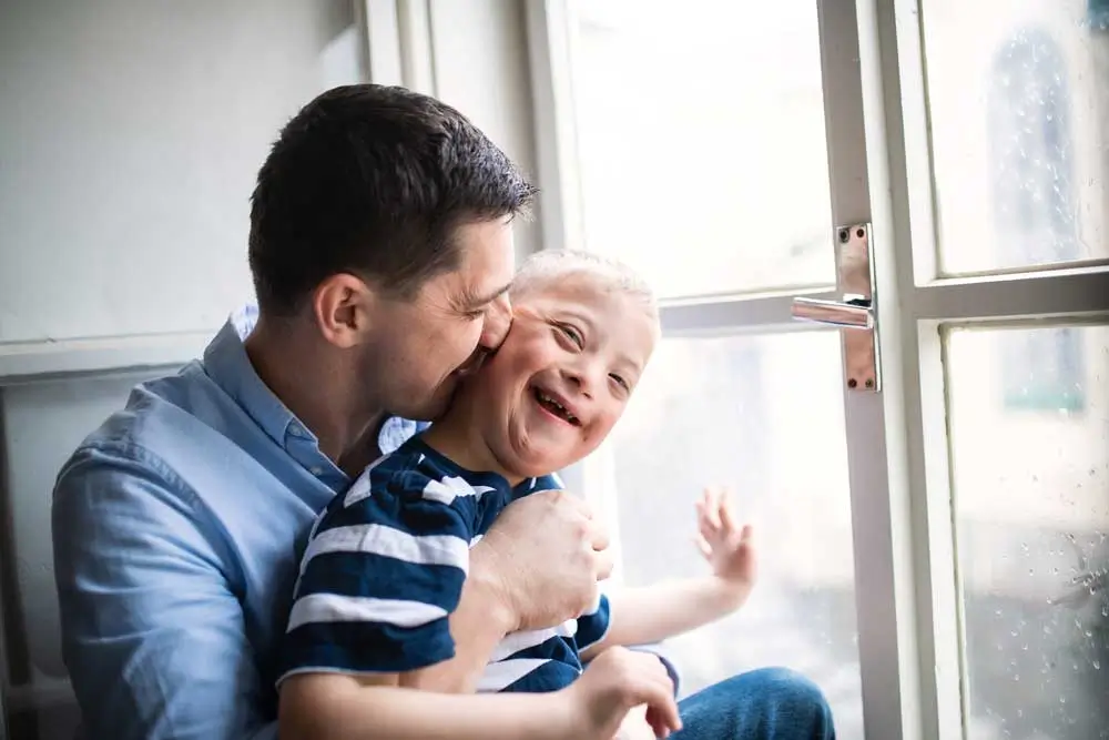 Father shares a tender moment with his young son with Down syndrome, both smiling and enjoying time together by the window.