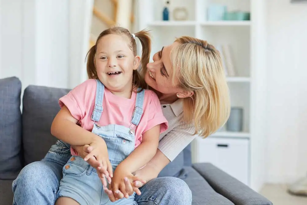 NDIS registered provider Melbourne helps a mother and her young daughter with Down syndrome share a joyful moment on the sofa, smiling and laughing together.