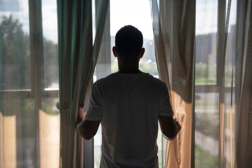 A man stands facing a window with curtains drawn, looking out into the distance. This image represents isolation, a common symptom when asking 'What is depression?' and how it affects people experiencing it.