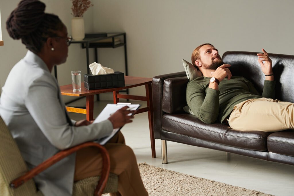 Therapist taking notes while a man lies on a couch, talking during a therapy session. Therapy is a crucial part of treatment when exploring 'What is depression?' and its impacts.