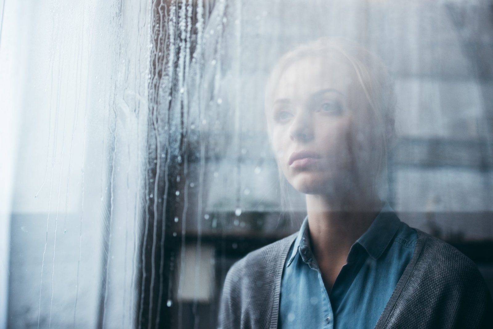 Woman staring through a glass window with a blank expression as raindrops run down the glass. This image captures the emotional pain associated with 'What is depression?' and its effects on mental health.