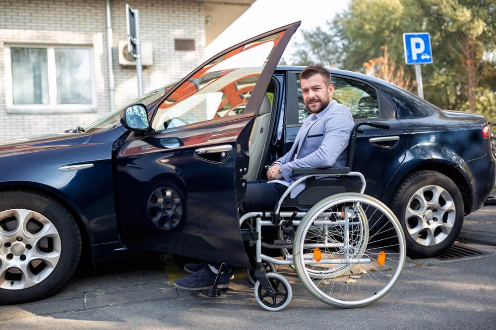 A person in a wheelchair in front of a black car, highlighting the importance of disability parking permits in providing accessible parking spaces for individuals with disabilities.