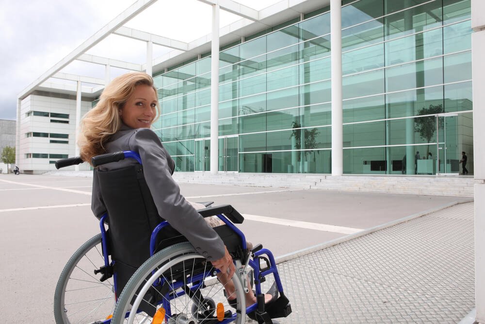 A person in a wheelchair going into a glass building, illustrating the role of disability parking permits in facilitating easy access to buildings and public spaces for wheelchair users.