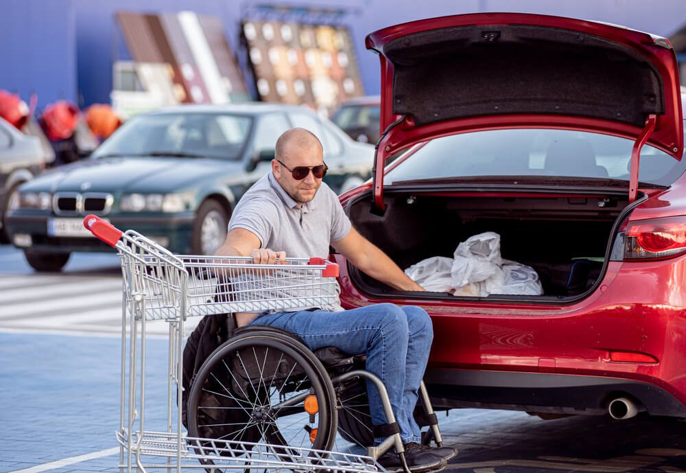 A person in a wheelchair with a shopping cart in front of a red car, emphasizing how disability parking permits help ensure convenient access to parking for people with disabilities while shopping.