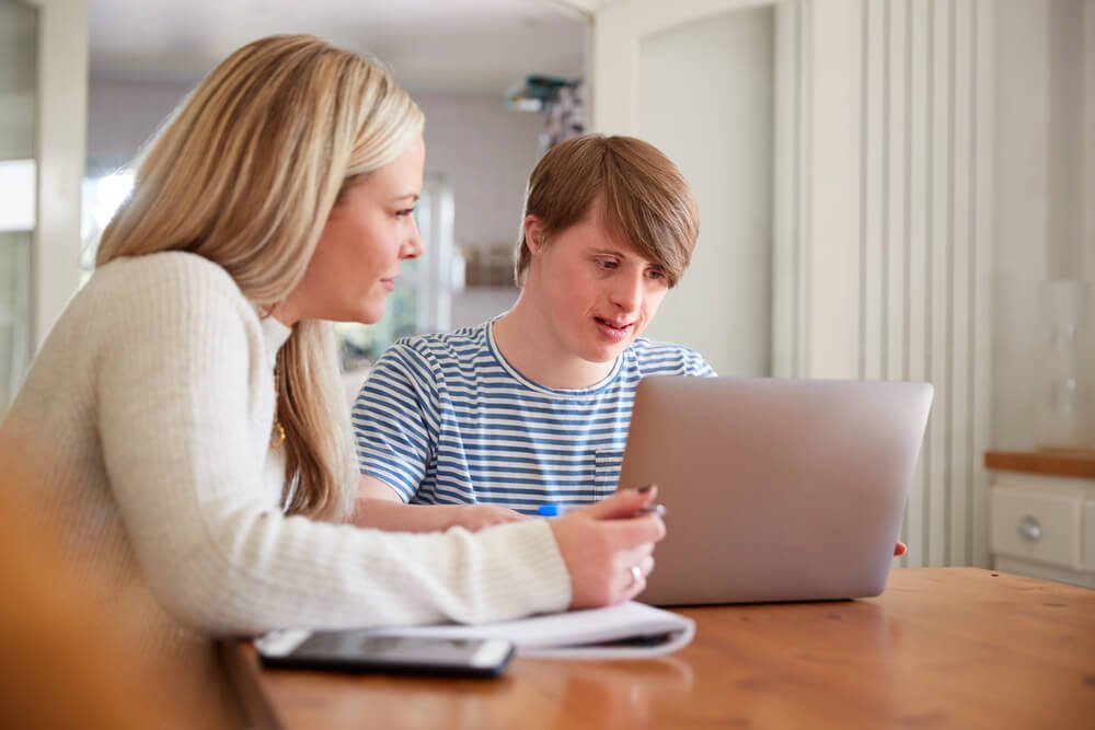 A young man with Down syndrome working on a laptop with the help of a support coordinator. They are sitting at a wooden table in a bright, cozy room. This scene highlights the importance of support coordinators in the NDIS framework, providing assistance and guidance.