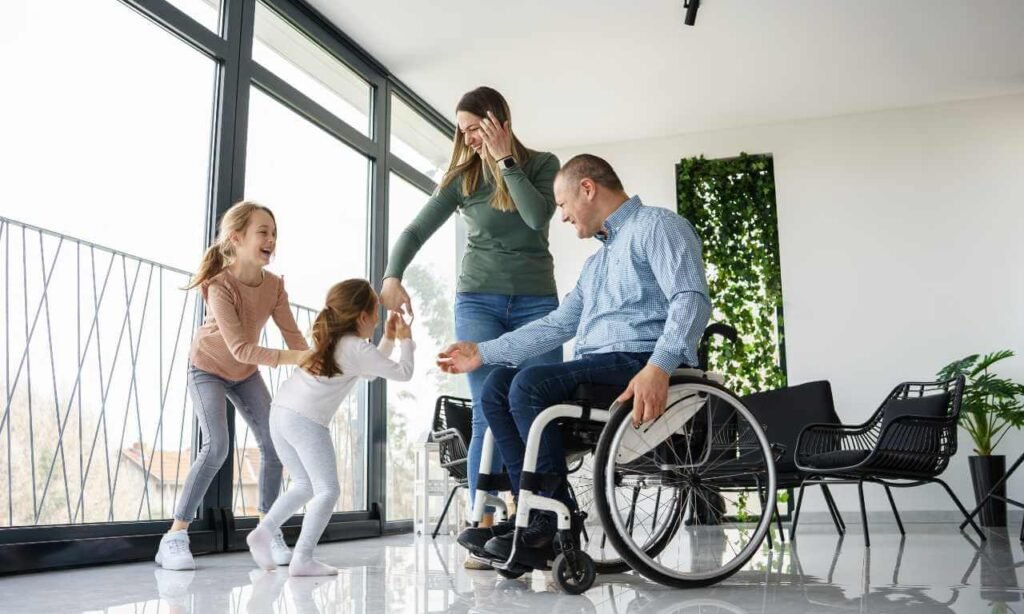 A physically challenged man in a wheelchair joyfully interacting with his family, including two young children and a woman, in a bright and airy living room. This image depicts personal care support in disability services, focusing on family inclusion and emotional well-being.