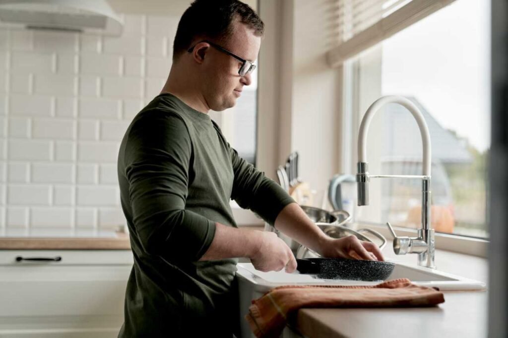 An adult man with Down syndrome wearing glasses, washing dishes in a bright kitchen. The scene showcases personal care support in disability services, promoting life skills and self-reliance.