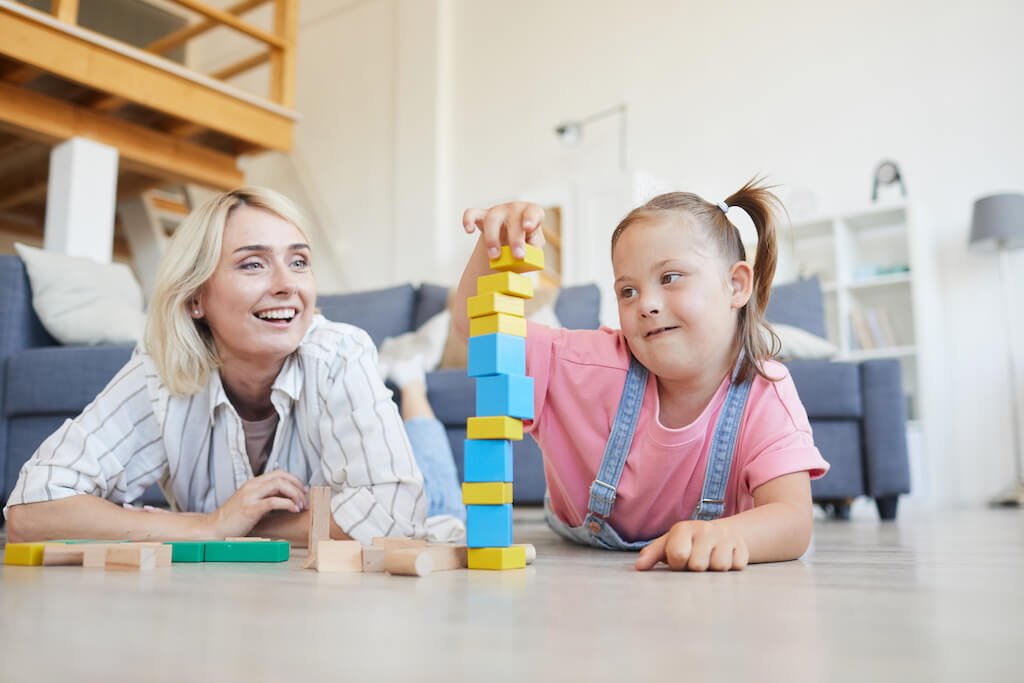 A young girl with Down syndrome is stacking colorful blocks while lying on the floor with her mother, illustrating an engaging and supportive interaction as part of NDIS support coordination in a bright, cozy living room.