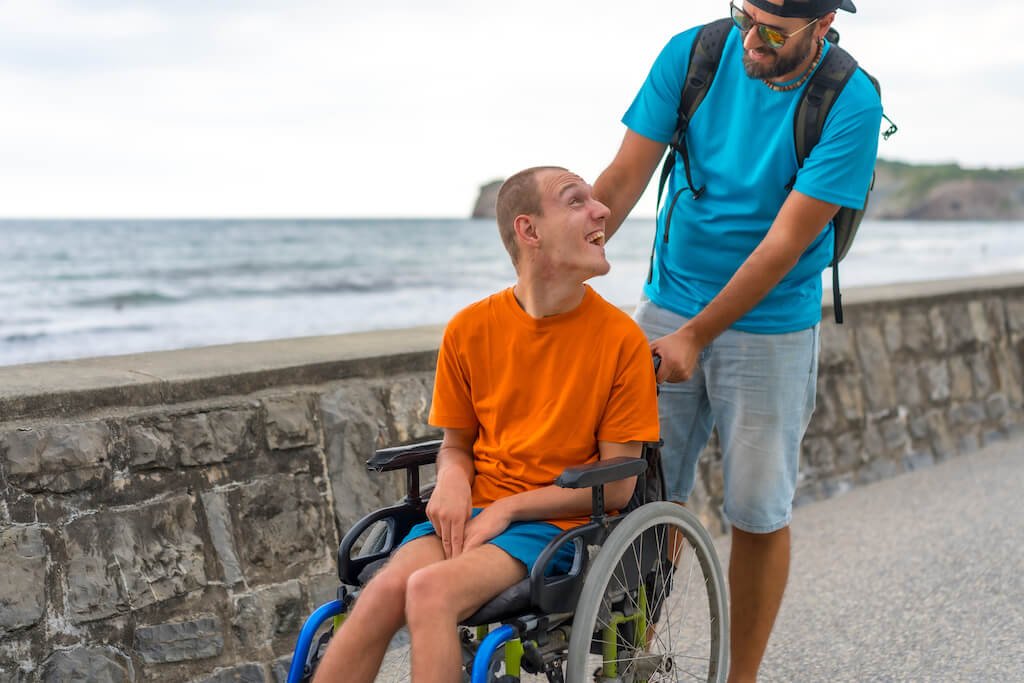 A person in a wheelchair is being pushed along a seaside promenade by a caregiver. Both individuals are smiling, enjoying the outdoor activity. The scene highlights daily activities NDIS, emphasizing the importance of support and inclusivity for individuals with disabilities in engaging with their environment.