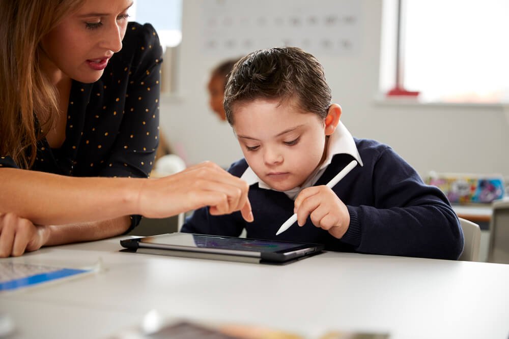 A young female NDIS support worker helps a child with Down syndrome use a tablet. The support worker is pointing at the screen, providing guidance and assistance. This image illustrates the role of an NDIS support worker in facilitating learning and technology use for children with disabilities.