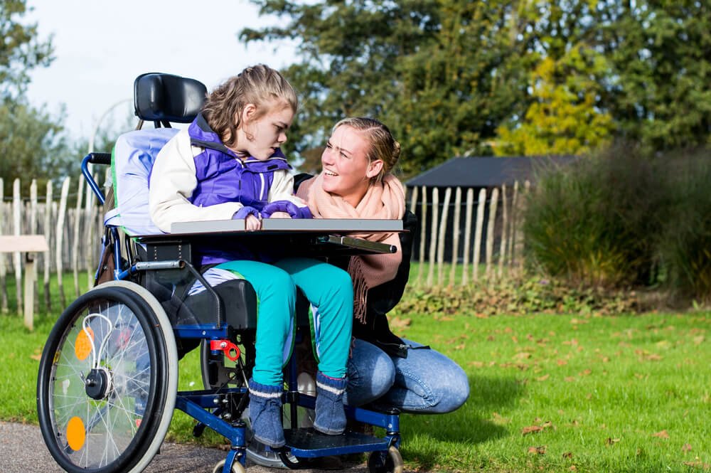 An NDIS support worker interacts with a child in a wheelchair outdoors. The support worker is kneeling beside the wheelchair, smiling and providing companionship. This image showcases the emotional and social support provided by NDIS workers to enhance the quality of life for children with disabilities.