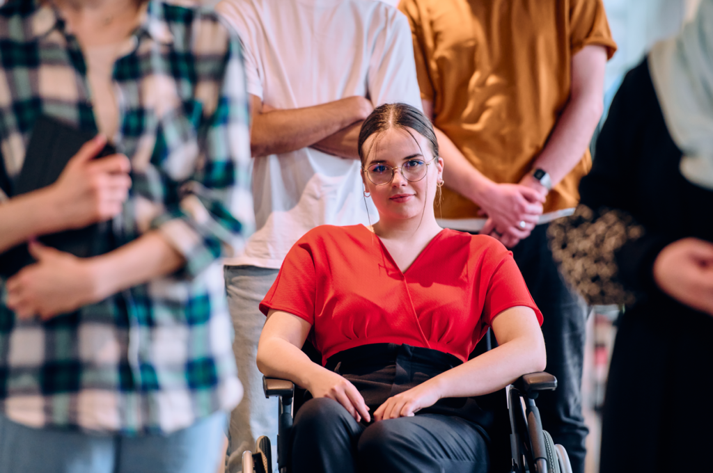 A woman in a wheelchair confidently faces the camera, surrounded by a diverse group, symbolising inclusivity and the support provided by NDIS service providers.