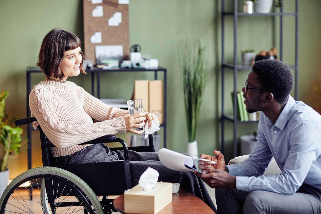 In the process to find a support worker NDIS, a woman in a wheelchair holds a glass of water while talking to a man taking notes in a professional office setting.