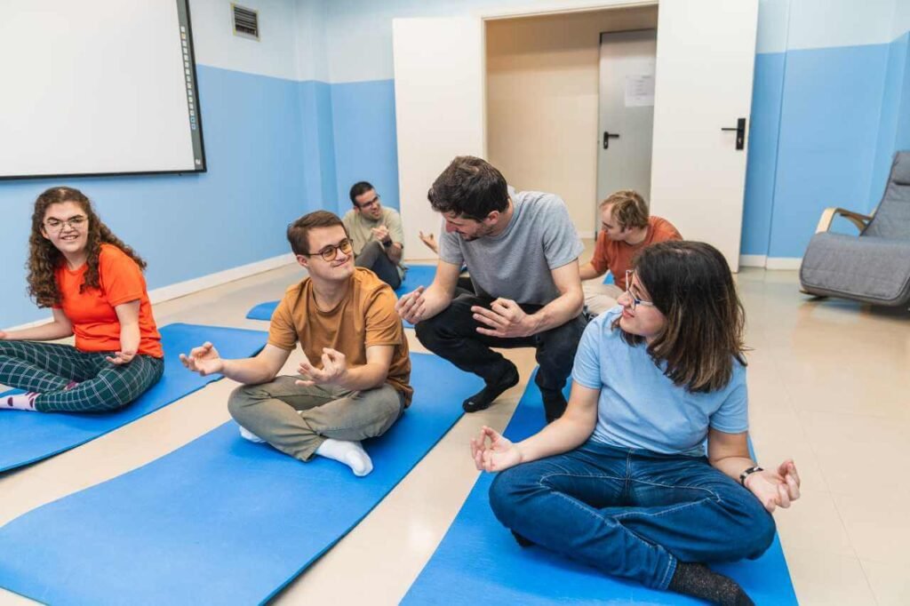 NDIS support worker instructs a group of individuals with Down syndrome during a yoga session. Participants are seated on yoga mats, engaging in a guided meditation or breathing exercise. This image highlights the involvement of NDIS support workers in promoting physical activity and mental well-being among people with disabilities.